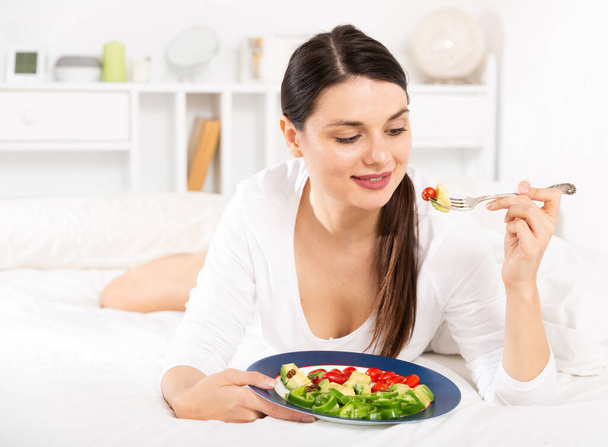 Pretty woman in white dress eating vegetable salad in bed - Foto, afbeelding