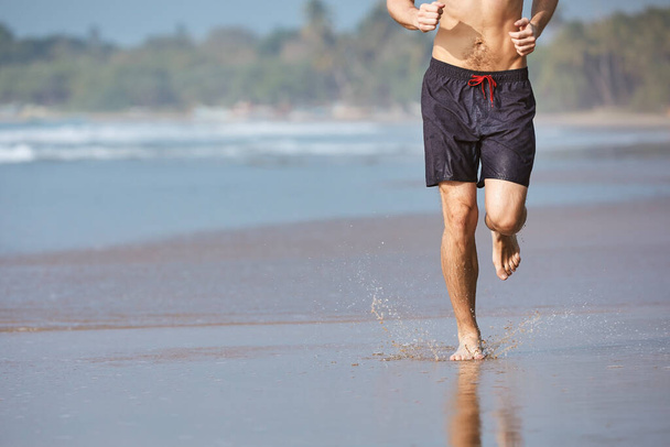 Man running on sand beach. Front view of barefoot runner against coastline. - Photo, image