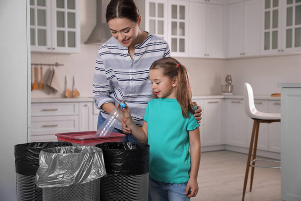 Young woman and her daughter throwing plastic bottle into trash bin in kitchen. Separate waste collection - Фото, зображення