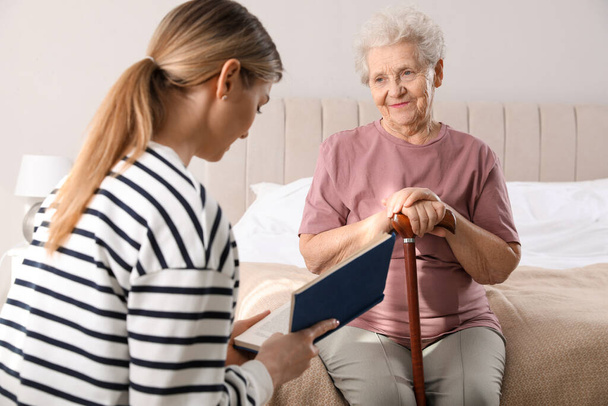 Young caregiver reading book to senior woman in bedroom. Home care service - Photo, Image