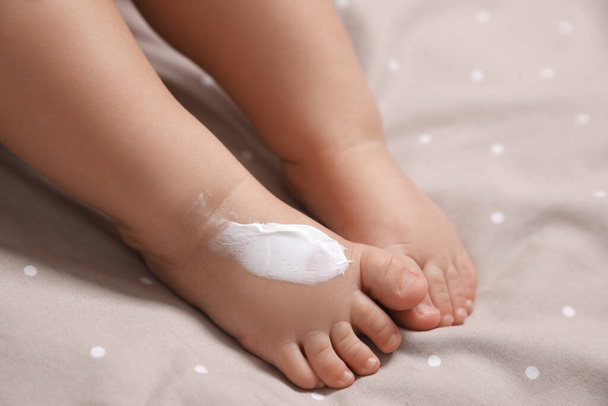 Cute little baby with moisturizing cream on her foot in bed, closeup - Fotó, kép