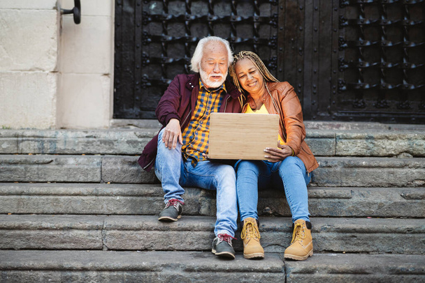 όμορφο ηλικιωμένο ζευγάρι ηλικιωμένων που χρησιμοποιεί τεχνολογία, υπολογιστή που κάθεται στις σκάλες της πόλης τη νύχτα. Ασιάτισσα Αφρικανή Αμερικανίδα. - Φωτογραφία, εικόνα