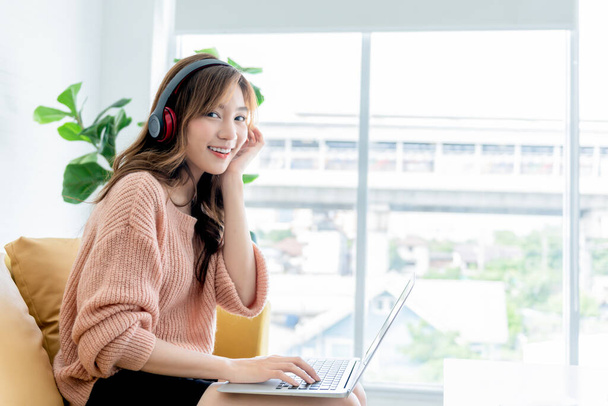 Portrety Azjatki, atrakcyjnej i młodej, siedzącej na kanapie, noszącej słuchawki i pracującej w domu, korzystającej z notebooków, do pracy w domu i w biznesie online - Zdjęcie, obraz