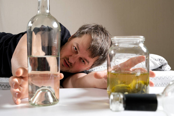 Un hombre borracho con una adicción al alcohol toma una botella de vodka y salmuera con encurtidos en vinagre. - Foto, Imagen