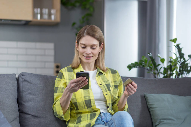 Happy woman shopping online sitting at home on the couch, using phone and bank credit card - Fotó, kép