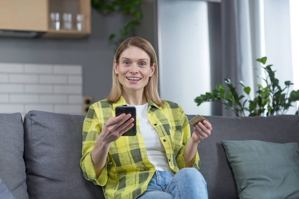 Portrait of a happy woman at home, housewife sitting on sofa happy and looking at camera smiling, holding phone and bank credit card for online shopping - 写真・画像