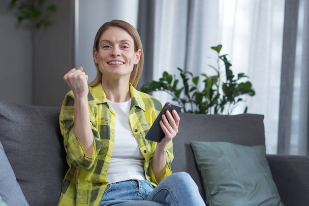 Happy woman at home, looking at camera and smiling rejoices and celebrates victory, sitting on sofa holding phone - Foto, Bild