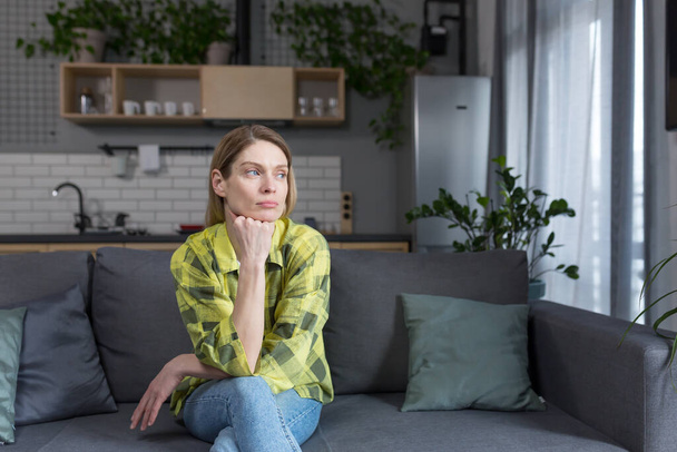 Pensive middle aged woman sitting at home on sofa thinking - 写真・画像