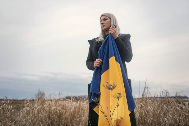 Russia's war against Ukraine. Young, beautiful patriot girl standing / running on a burnt wheat field with a flag. Grainy emotional picture, pain and despair of the people of an independent country. - Фото, зображення