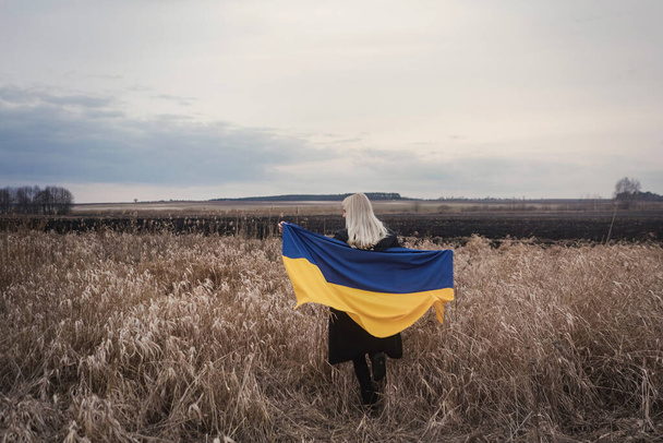 Ruská válka proti Ukrajině. Mladá, krásná vlastenecká dívka stojící / běhající na spáleném pšeničném poli s vlajkou. Zrnitý emocionální obraz, bolest a zoufalství lidí z nezávislé země. - Fotografie, Obrázek