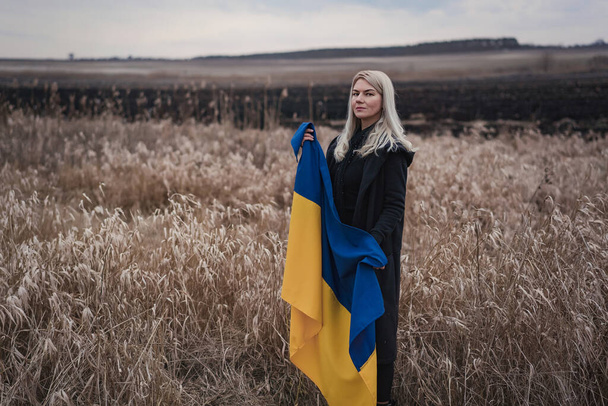 Russia's war against Ukraine. Young, beautiful patriot girl standing / running on a burnt wheat field with a flag. Grainy emotional picture, pain and despair of the people of an independent country. - Photo, Image