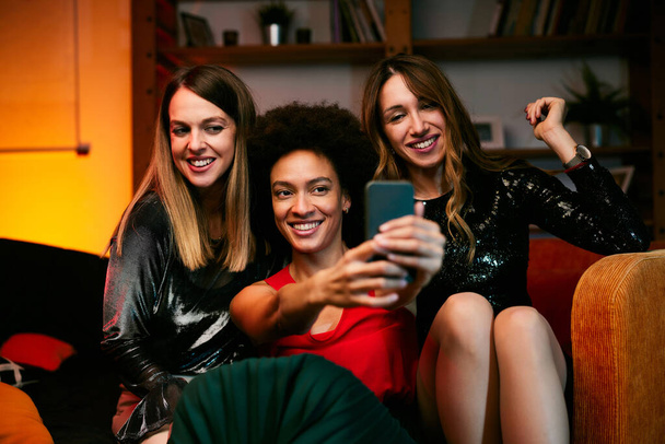 Üç çok kültürlü bayan arkadaş akşamları oturma odasında oturup ev partisinde selfie çekiyorlar.. - Fotoğraf, Görsel