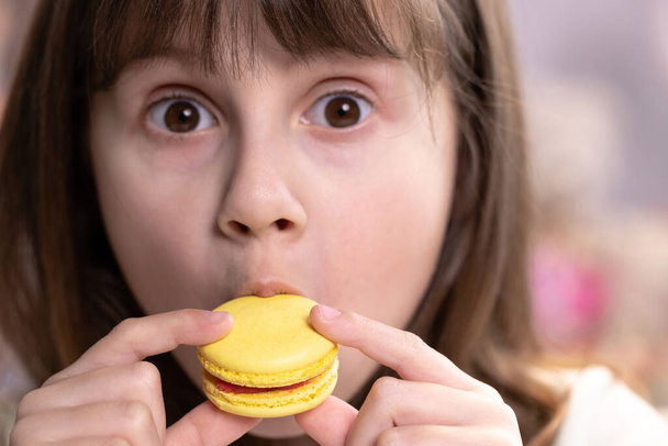 School girl on a home background, licks her lips, eating one yellow macaron. Dessert person, sweet tooth, gourmet. Feeling good, delicious - Фото, изображение