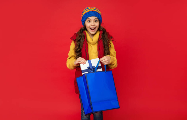 изумленная девушка-подросток в трикотажной шляпе держать торговый мешок и подарить коробку на красном фоне, покупка - Фото, изображение