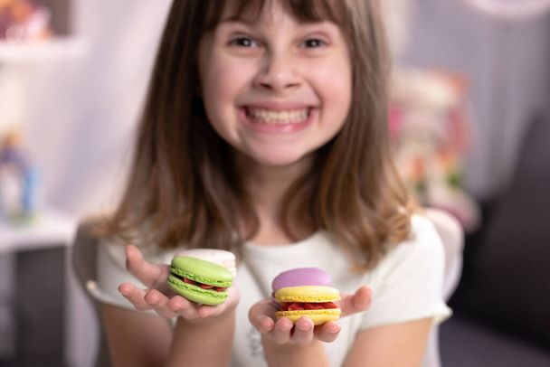 Smiling cute school kid girl holds four tasty cookies in her hands and look at camera. Traditional French multicolored macaroon. Food concept - Фото, изображение