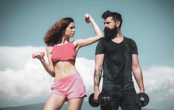 Sporty couple working out with dumbbells on sky background. Healthy lifestyle concept. Beautiful attractive fitness friends. Abs fitness woman body. Female model in sportswear exercising outdoors. - 写真・画像