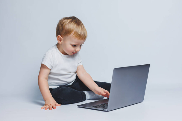 Un petit garçon de 3-4 ans est assis avec un ordinateur portable sur un fond blanc. Un enfant portant un T-shirt blanc et un pantalon noir regarde un ordinateur portable. Enfants modernes - Photo, image