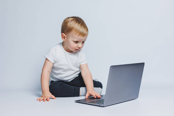 Mały chłopiec w wieku 3-4 lat siedzi z laptopem na białym tle. Dziecko w białej koszulce i czarnych spodniach patrzy na laptopa. Nowoczesne dzieci - Zdjęcie, obraz