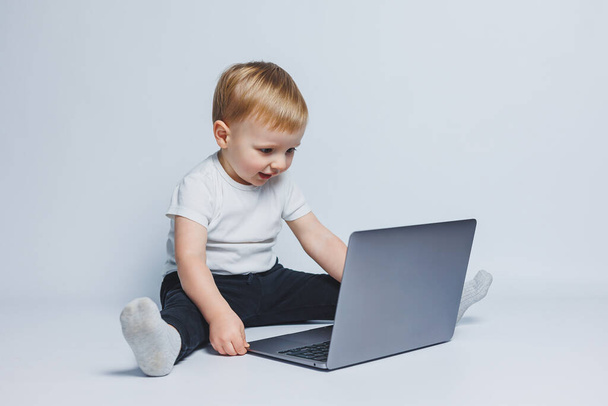 Un niño de 3-4 años se sienta con un portátil sobre un fondo blanco. Un niño con una camiseta blanca y un pantalón negro mira un portátil. Niños modernos - Foto, Imagen