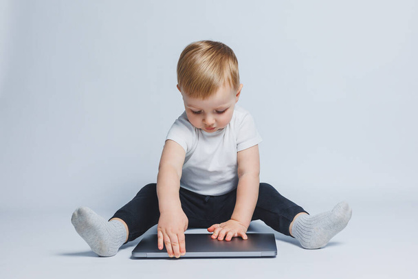 Μικρό έξυπνο αγόρι 3-4 ετών κάθεται με ένα φορητό υπολογιστή σε λευκό φόντο. Ένα παιδί με λευκό μπλουζάκι και μαύρο παντελόνι κάθεται σε ένα φορητό υπολογιστή και κοιτάζει την οθόνη. Σύγχρονα προοδευτικά παιδιά - Φωτογραφία, εικόνα