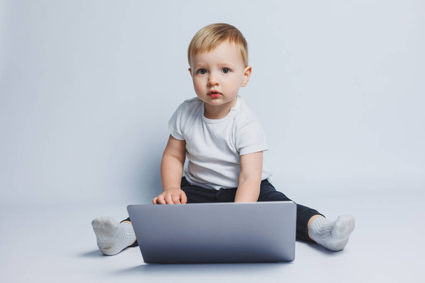 Um garoto inteligente de 3-4 anos se senta com um laptop em um fundo branco. Uma criança com uma camiseta branca e calças pretas senta-se em um laptop e olha para a câmera. Crianças modernas - Foto, Imagem