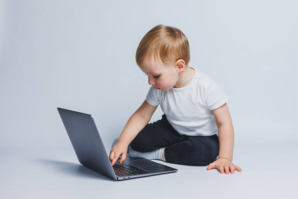 Pequeño niño inteligente de 3-4 años de edad se sienta con una computadora portátil sobre un fondo blanco. Un niño con una camiseta blanca y un pantalón negro se sienta en una computadora portátil y mira a la pantalla. Niños progresivos modernos - Foto, Imagen