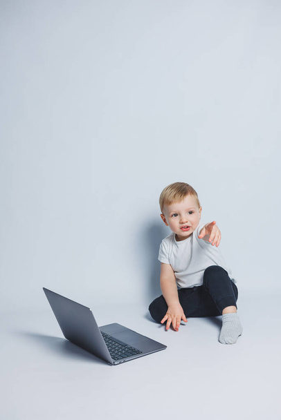 Akıllı bir çocuk 3-4 yaşlarında beyaz bir arka planda dizüstü bilgisayarla oturuyor. Beyaz tişörtlü ve siyah pantolonlu bir çocuk dizüstü bilgisayarın başına oturur ve kameraya bakar. Modern çocuklar - Fotoğraf, Görsel