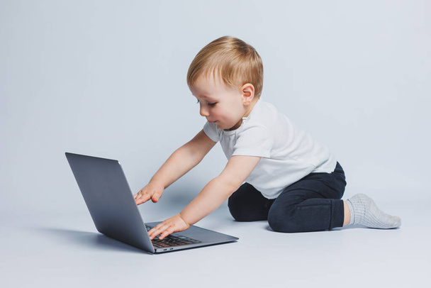 3-4 yaşlarında küçük zeki bir çocuk beyaz bir arka planda dizüstü bilgisayarla oturuyor. Beyaz tişörtlü ve siyah pantolonlu bir çocuk dizüstü bilgisayarın başına oturur ve ekrana bakar. Modern yenilikçi çocuklar - Fotoğraf, Görsel