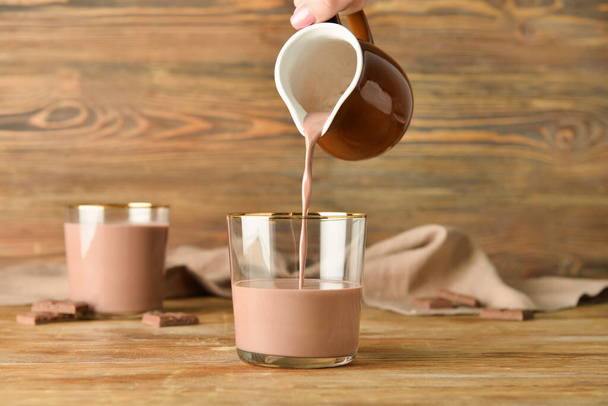 Pouring of delicious chocolate milk from jug into glass on wooden background - Photo, Image