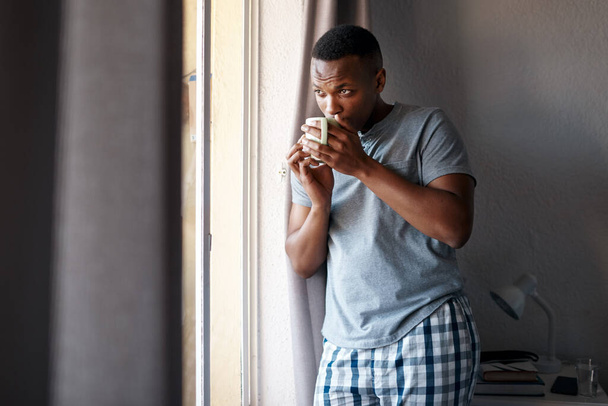 Theres nothing like coffee to get me through the day. Cropped shot of a handsome young man standing and looking contemplative in his bedroom while enjoying a cup of coffee. - Foto, Imagem
