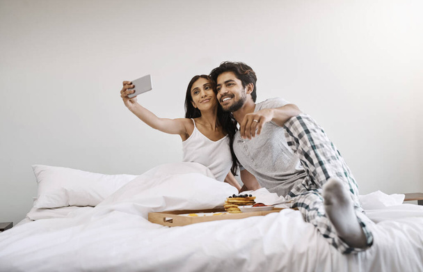We woke up like this. Full length shot of an affectionate young couple taking selfies while having breakfast in bed. - Photo, Image