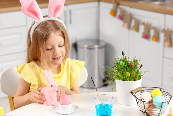 Adorable little girl with figurine of bunny painting Easter eggs in kitchen at home - Valokuva, kuva