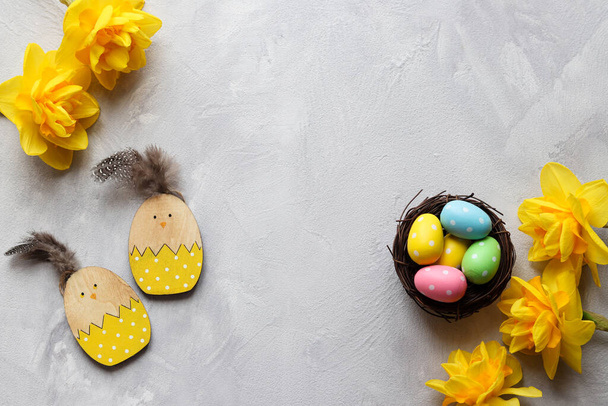 Easter composition with yellow daffodils, wooden chicken toys and nest with colorful eggs against grey textured background with copy space. Top view. - 写真・画像