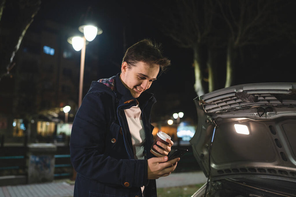 Ένας ευτυχισμένος νεαρός ενήλικας καυκάσιος στέκεται δίπλα στο όχημά του με ανοιχτή κουκούλα και χαλασμένη χαλασμένη μηχανή χαμογελώντας ανέμελα κρατήστε το τηλέφωνο καλώντας την υπηρεσία ρυμούλκησης για βοήθεια στην ιδέα οδικής βοήθειας - Φωτογραφία, εικόνα