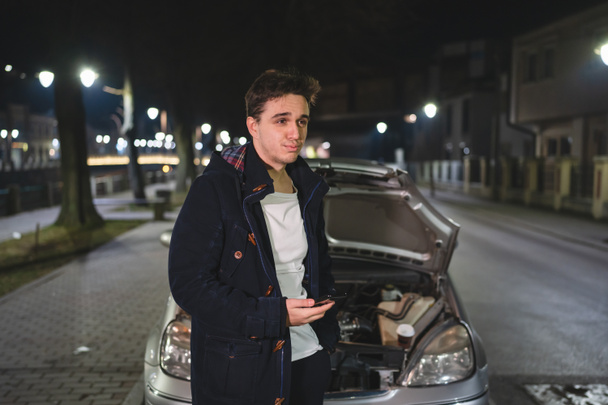 Jeden mladý dospělý kavkazský muž stojící u svého vozidla s otevřenou kapotou a rozbitým motorem držící telefon volající odtahovou službu o pomoc na silnici Silniční asistenční koncept v tmavé noci - Fotografie, Obrázek