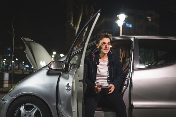 Ένας νεαρός ενήλικας καυκάσιος κάθεται στο όχημά του με ανοιχτή πόρτα και κουκούλα και χαλασμένη μηχανή κρατώντας ένα τηλέφωνο καλώντας την υπηρεσία ρυμούλκησης για βοήθεια στο δρόμο έννοια οδικής βοήθειας σε σκοτεινή νύχτα - Φωτογραφία, εικόνα