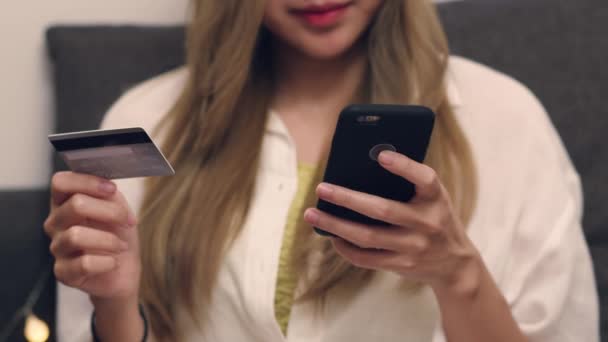 Close Up Azjatki w białej koszuli siedzi na kanapie i za pomocą smartfona przeglądania mediów społecznościowych i zakupu płatności online na karcie kredytowej podczas pobytu w salonie. - Materiał filmowy, wideo
