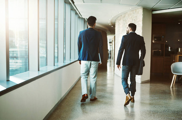 Success doesnt happen until people get moving. Rearview shot of two young businessmen walking down a corridor of a modern office. - Fotó, kép