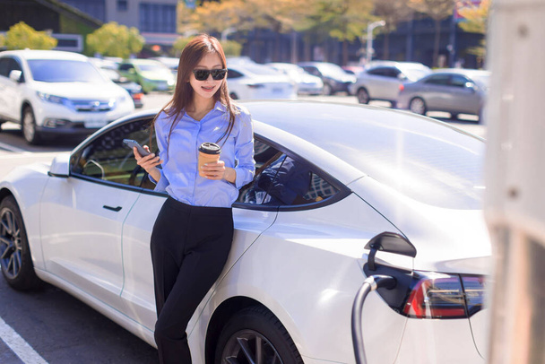 Business  woman standing on city parking near electric car, charging automobile battery from small city station, drinking coffee and using smartphone - Photo, Image