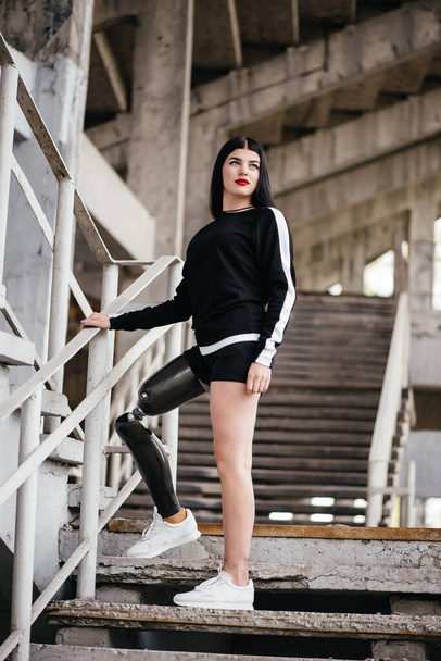 Photo of healthy disabled woman in sportswear with prosthesis leg standing at the stairs and looking aside outdoor - Zdjęcie, obraz