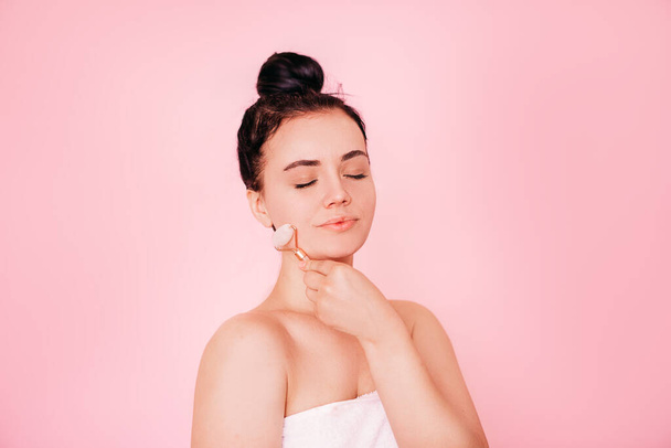 Une jeune femme détendue debout isolée sur un fond rose et souriante les yeux fermés, à l'aide d'un rouleau facial en quartz rose. le concept de soins à domicile - Photo, image