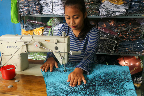 Krawcowa azjatycka szyje ubrania na maszynie do szycia. Uśmiechnięta krawcowa w warsztacie. Młoda krawcowa pracująca w fabryce tekstyliów - Zdjęcie, obraz