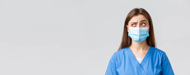 Covid-19, πρόληψη του ιού, εργαζόμενοι στον τομέα της υγειονομικής περίθαλψης και καραντίνα έννοια. Σκεπτική γυναίκα νοσοκόμα ή γιατρός με μπλε ποδιά, ιατρική μάσκα, σηκώστε το ένα φρύδι με περιέργεια και σύγχυση, κοιτάξτε στην πάνω αριστερή γωνία - Φωτογραφία, εικόνα
