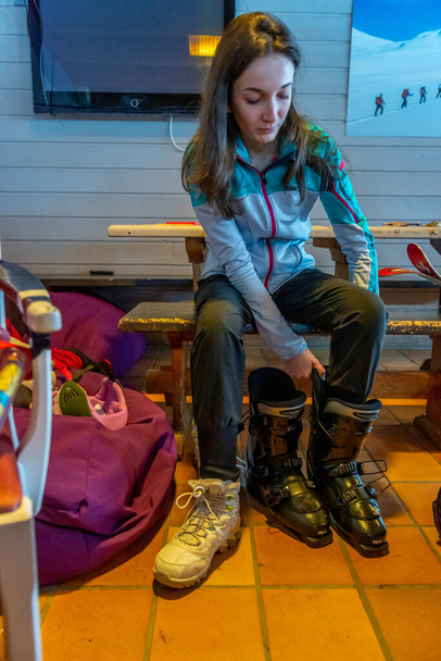 Woman with brown hair preparing for skiing, looking down while trying on one black ski shoes inside a building, vertical - Photo, Image