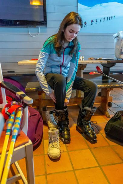 Mulher com cabelo castanho se preparando para esquiar, olhando para baixo enquanto experimenta seus sapatos de esqui pretos dentro de um prédio, vestindo dois sapatos, bastões de esqui na frente, vertical - Foto, Imagem