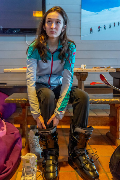 Woman with brown hair preparing for skiing, looking up while trying on her black ski shoes inside a building, vertical - Photo, Image
