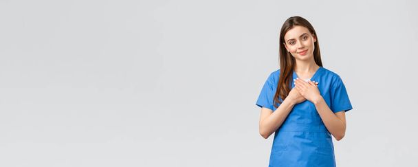 Healthcare workers, prevent virus, insurance and medicine concept. Touched and flattered female nurse or doctor in blue scrubs, tilt head and touch heart, smile from compliment, nice gesture - Photo, Image
