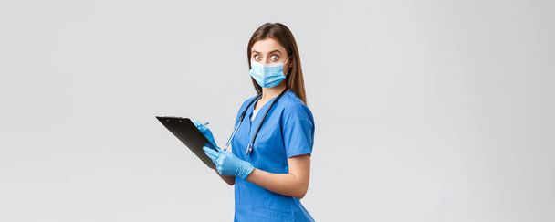 Covid-19, preventing virus, health, healthcare workers and quarantine concept. Shocked and startled female nurse or doctor in blue scrubs, medical mask, holding clipboard stare surprised camera - Foto, afbeelding