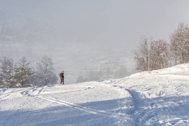 Mujer de cabello castaño, con ropa de esquí negro y casco esquiando por la cima de una montaña, mirando hacia atrás en dirección de la cámara, vista trasera, hermoso paisaje de nieve, valle en la distancia, niebla, horizontal - Foto, imagen