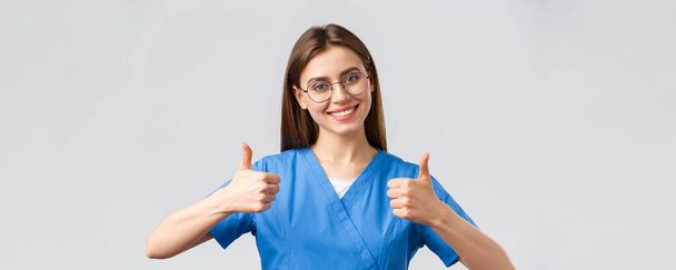 Healthcare workers, medicine, insurance and covid-19 pandemic concept. Excited happy female nurse in blue scrubs, doctor show thumbs-up in support or approval, encourage make tests in clinic - Foto, imagen